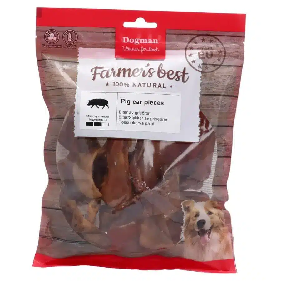 Ett rött och genomskinligt plastpaket med Dogman Farmer's Best Pig Ear Pieces hundgodis. Förpackningen har en bild på en glad hund i det nedre högra hörnet och en etikett som anger att produkten är 100 % naturlig med tuggstyrkedetaljer.